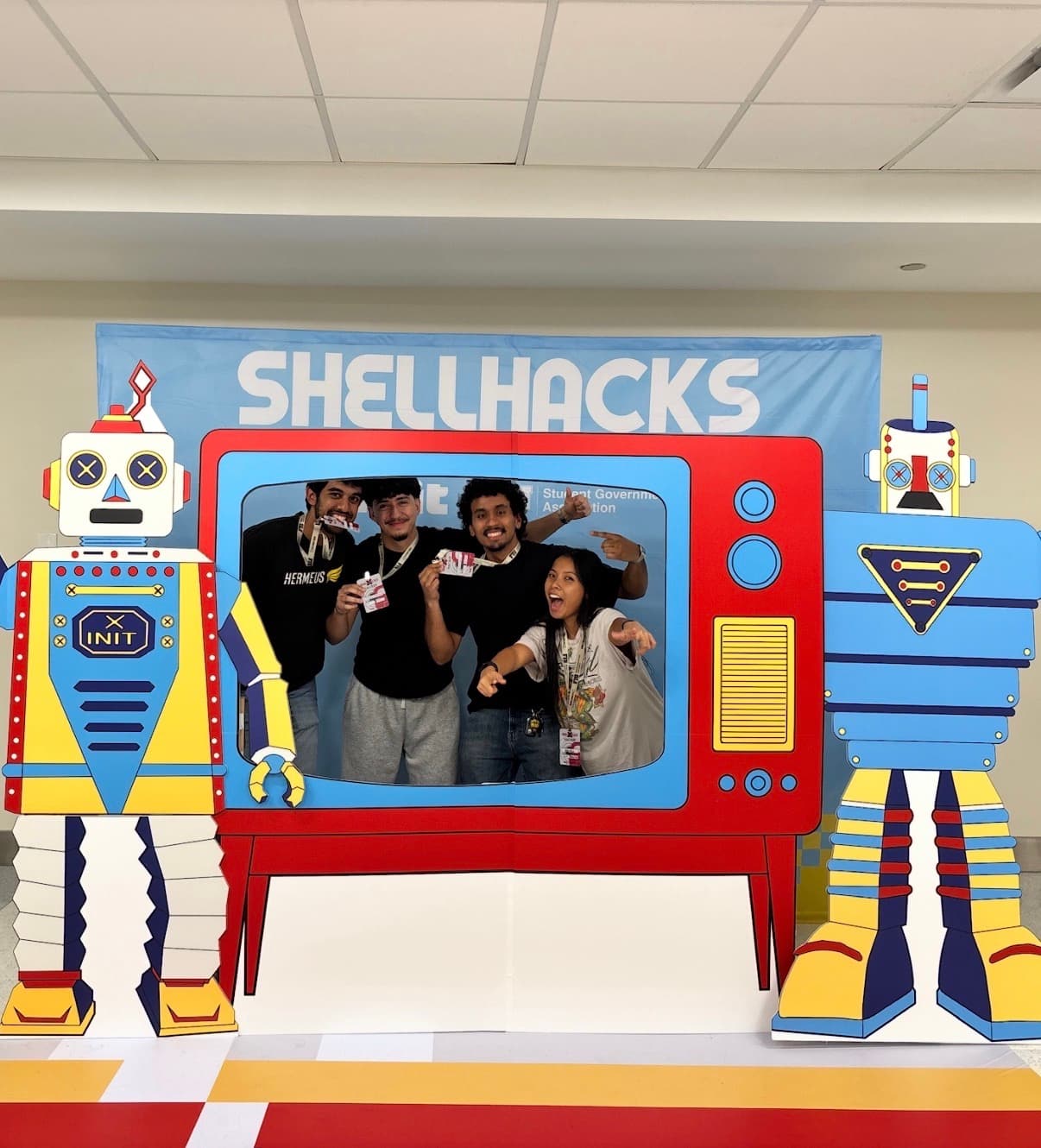 Four participants posing inside a colorful television frame beside robot cutouts at ShellHacks.